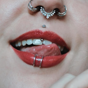 May include: Close-up of a person's mouth with red lipstick and multiple piercings. The person is sticking their tongue out and has a silver ring piercing on their lower lip, a silver ring piercing on their nose, and a silver stud piercing on their upper lip.