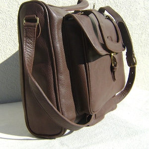 May include: A brown leather briefcase with a long strap and a buckle closure. The bag has a flap with a buckle closure and a smaller pocket on the front.