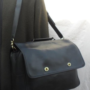 Może przedstawiać: Czarna skórzana teczka z klapką i dwoma złotymi guzikami. Teczka ma długi, regulowany pasek na ramię.