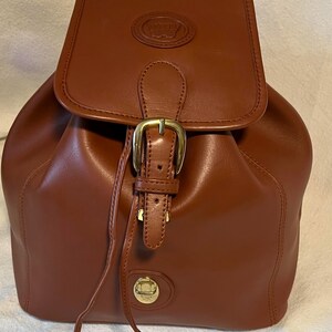 May include: A brown leather backpack with a gold buckle and a circular logo on the front. The backpack has a drawstring closure and a flap with a buckle.