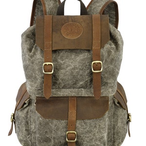 May include: A gray canvas backpack with brown leather accents and buckles. The backpack has a large main compartment and a smaller front pocket. The front pocket has a brown leather flap with a logo that says "Lord & Buddy" in a circle.