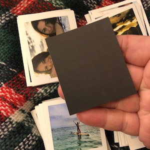 May include: A dark gray square magnet. The magnet is being held in a hand.