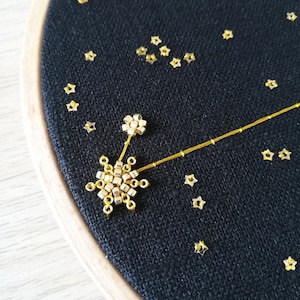 May include: A black fabric hoop with gold star-shaped sequins and a gold beaded constellation design. The constellation is made of small gold beads and a thin gold thread. The text "Oph&eacute;lie Trichereau" is visible at the bottom of the hoop.