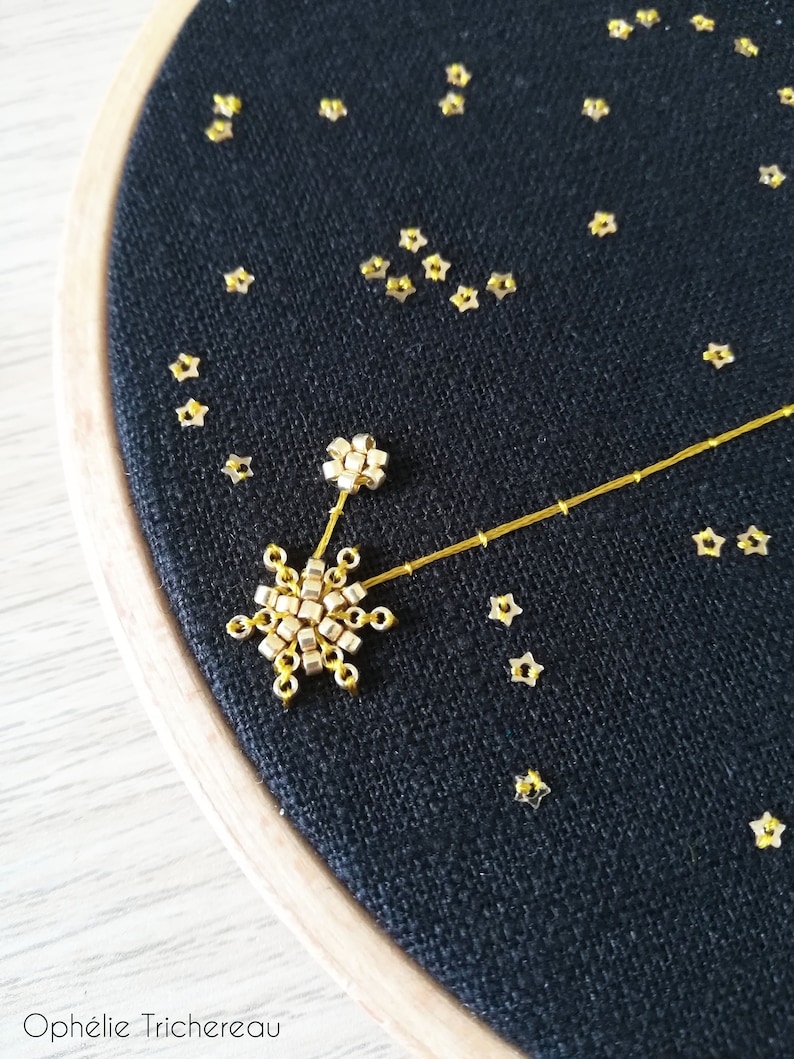 May include: A black fabric hoop with gold star-shaped sequins and a gold beaded constellation design. The constellation is made of small gold beads and a thin gold thread. The text "Oph&eacute;lie Trichereau" is visible at the bottom of the hoop.