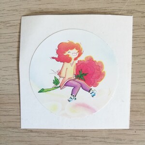 Peut inclure: Un autocollant représentant une illustration de dessin animé d'une fille aux cheveux roux assise sur une grande fleur rose. La fille sourit et porte une chemise jaune et un pantalon violet.