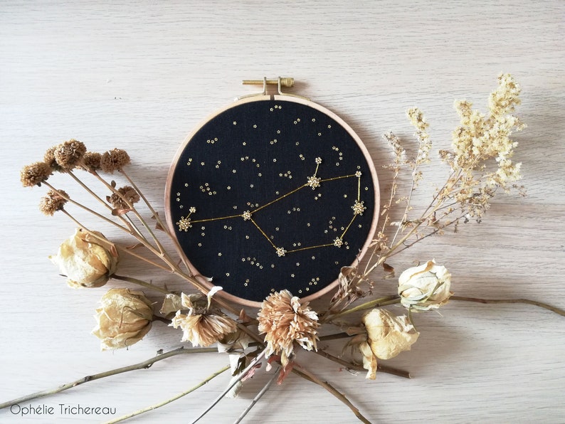 May include: A black embroidery hoop featuring a gold constellation design on a black fabric background. The hoop is surrounded by dried flowers.