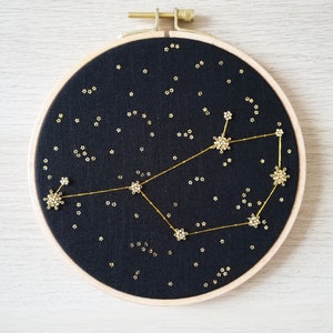 May include: A black embroidery hoop featuring a gold constellation design. The constellation is made up of small gold stars and a larger gold star design. The background is black fabric.