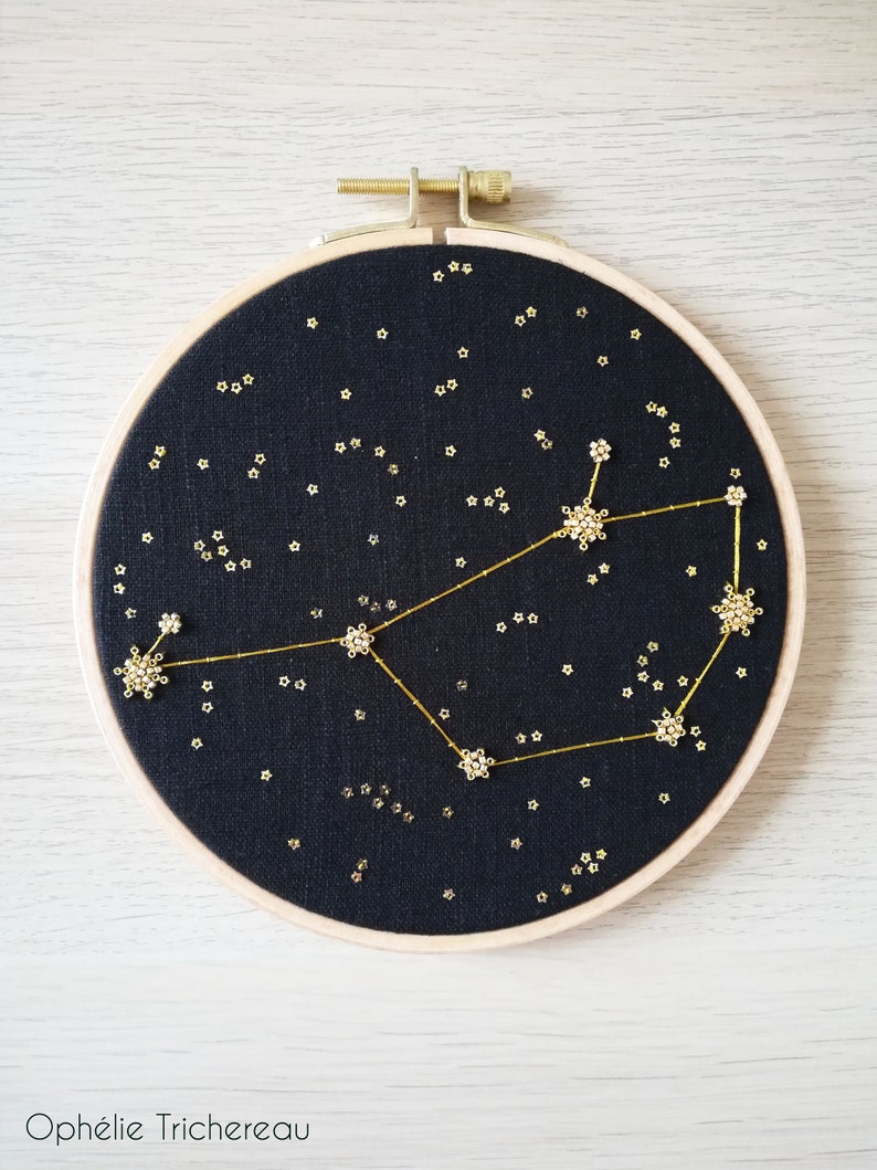 May include: A black embroidery hoop featuring a gold constellation design. The constellation is made up of small gold stars and a larger gold star design. The background is black fabric.