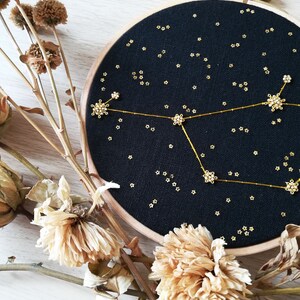 May include: A black embroidery hoop featuring a gold constellation design. The constellation is made of small gold beads and thread. The hoop is surrounded by dried flowers.