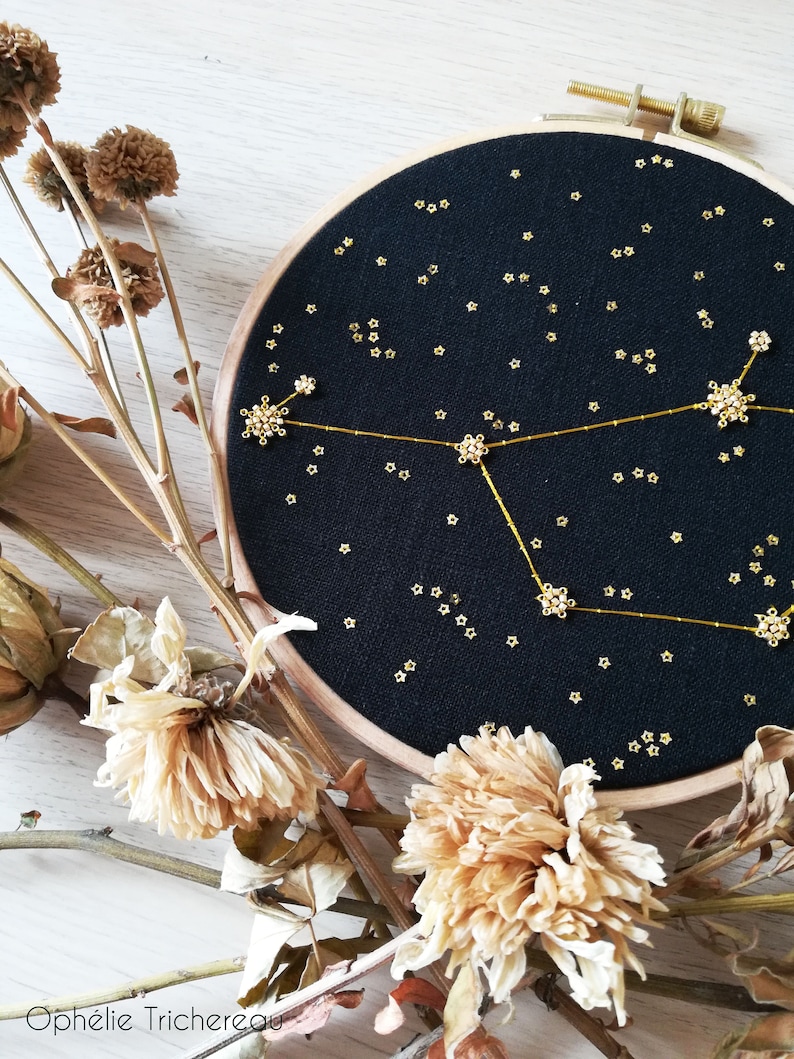 May include: A black embroidery hoop featuring a gold constellation design. The constellation is made of small gold beads and thread. The hoop is surrounded by dried flowers.