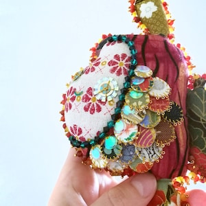 May include: A handmade fabric ornament with a red, white, and green color scheme. The ornament is decorated with beads, sequins, and fabric scraps. It has a whimsical, folk art style.