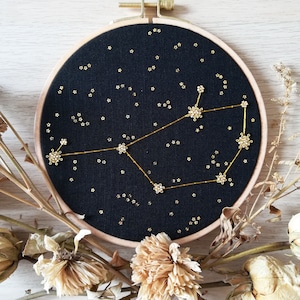 May include: A black embroidery hoop featuring a gold constellation design. The constellation is made of small gold beads and the background is a dark black fabric. The hoop is surrounded by dried flowers.