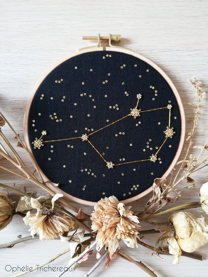 May include: A black embroidery hoop featuring a gold constellation design. The constellation is made of small gold beads and the background is a dark black fabric. The hoop is surrounded by dried flowers.