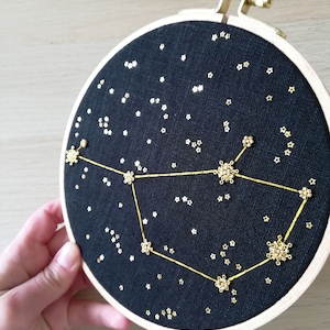 May include: A black embroidery hoop featuring a gold constellation design. The constellation is made of small gold beads and thread. The background is a dark blue fabric with small gold stars.