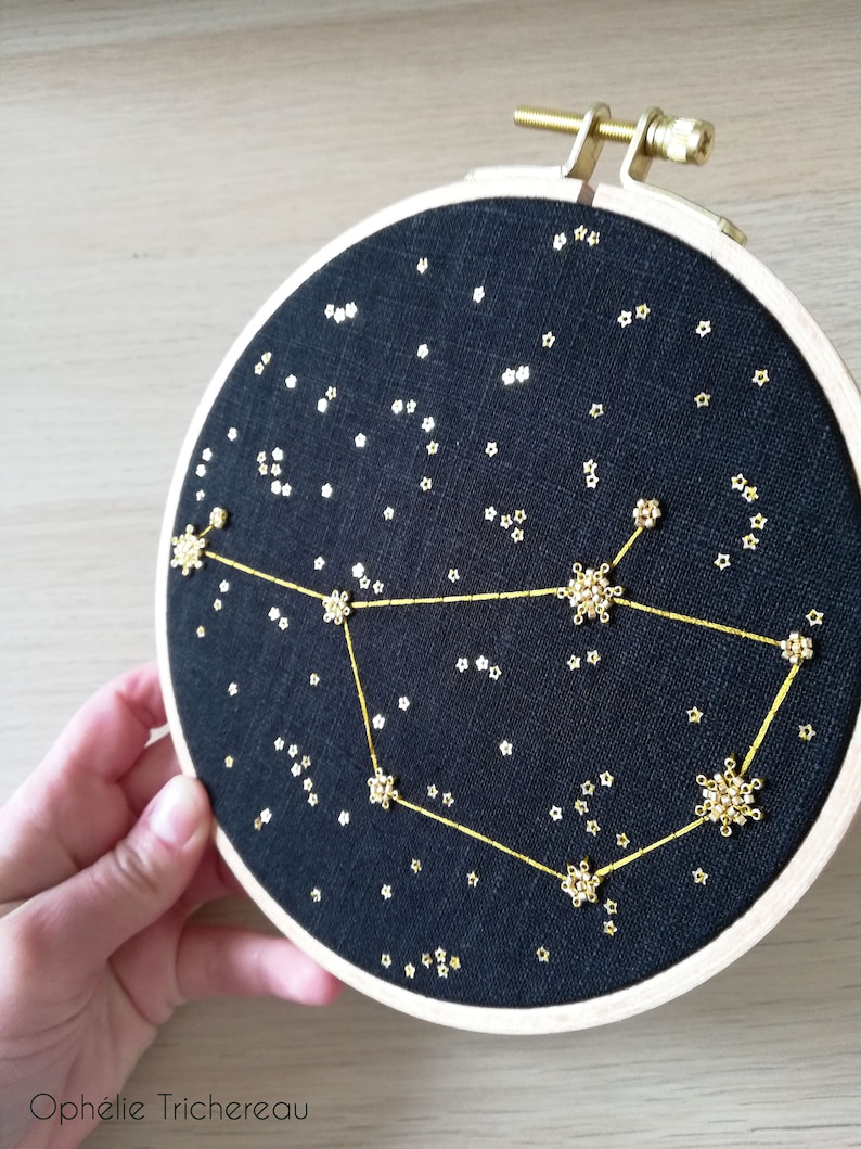 May include: A black embroidery hoop featuring a gold constellation design. The constellation is made of small gold beads and thread. The background is a dark blue fabric with small gold stars.