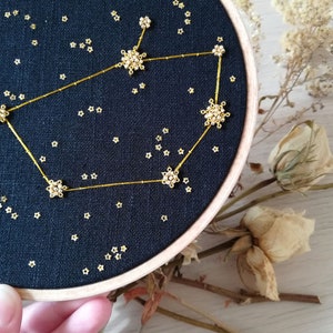May include: A black embroidery hoop featuring a gold constellation design. The constellation is made of small gold beads and thread. The background is a dark blue fabric with small gold stars.