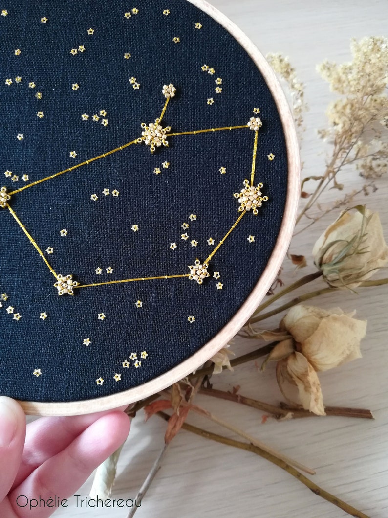May include: A black embroidery hoop featuring a gold constellation design. The constellation is made of small gold beads and thread. The background is a dark blue fabric with small gold stars.