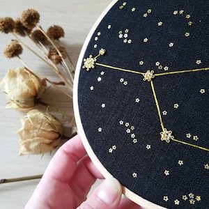 May include: A black embroidery hoop featuring a gold constellation design. The constellation is outlined in gold thread and the stars are made of gold beads. The hoop is held in a hand.