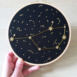 May include: A black embroidery hoop featuring a gold constellation design. The constellation is outlined with gold thread and the stars are made of gold beads. The hoop is on a light wood surface.