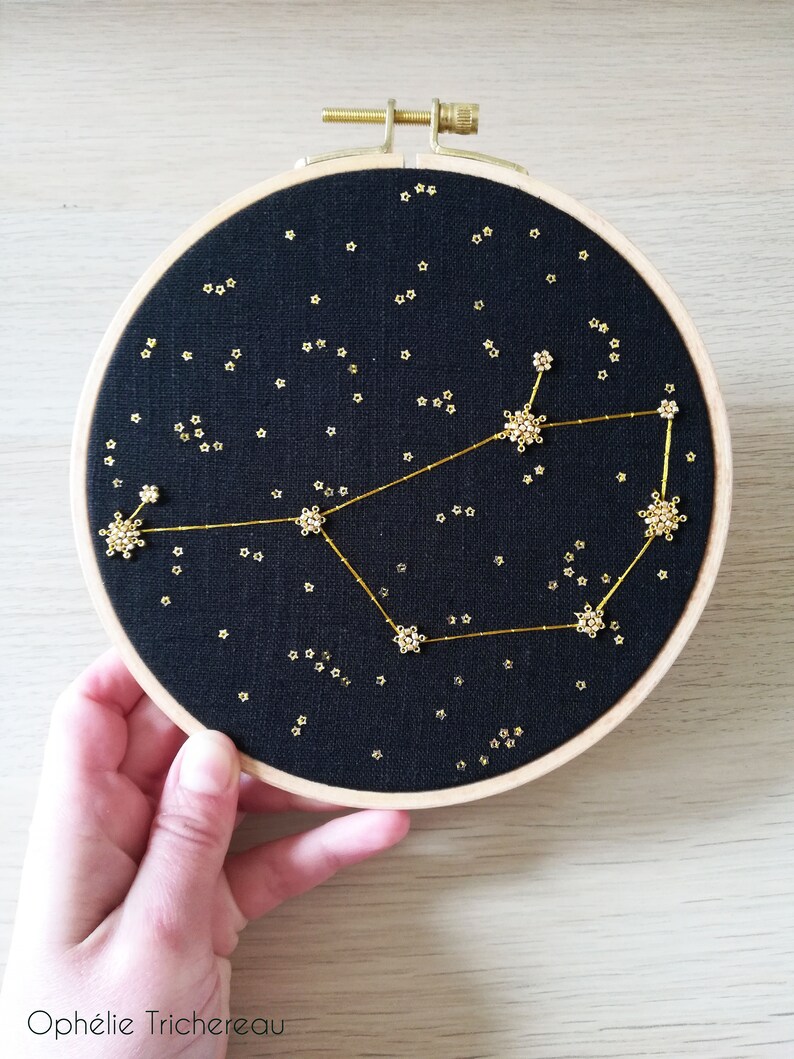 May include: A black embroidery hoop featuring a gold constellation design. The constellation is outlined with gold thread and the stars are made of gold beads. The hoop is on a light wood surface.