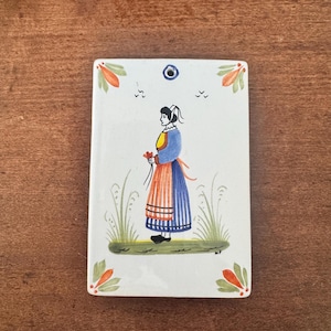 May include: Rectangular ceramic tile with a hand-painted illustration of a woman in traditional dress. The woman is holding flowers, standing in a field of grass. The tile has a hole at the top for hanging, with floral accents.