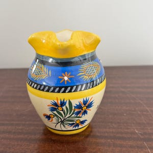 Puede incluir: Una pequeña jarra de cerámica pintada a mano con borde y asa amarillos. El cuerpo es de color crema con diseños florales y geométricos azules y amarillos. La jarra tiene una forma decorativa única.