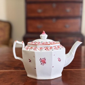 Könnte beinhalten: Weiße Porzellan-Teekanne mit achteckigem Korpus und Deckel. Die Teekanne ist mit roten und rosafarbenen Blumendesigns und einem zarten Blumenmuster am Rand verziert.