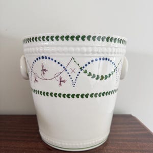 Könnte beinhalten: Ein weißer Keramik-Pflanztopf mit dekorativem Design. Der Pflanztopf hat grüne und blaue Blumenmuster mit kleinen Griffen auf beiden Seiten. Der Rand und der Boden sind grün eingefasst. Der Pflanztopf steht auf einer dunkelbraunen Holzoberfläche.