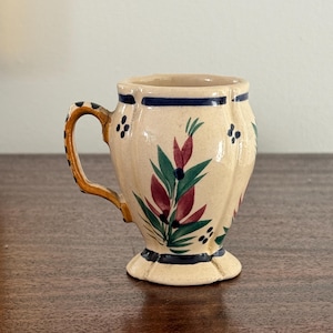 May include: A small, cream-colored ceramic mug with a handle. The mug features a floral design in red, green, and blue. The handle is yellow with brown accents. The mug has a blue stripe around the top and bottom.