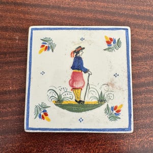 May include: Square ceramic tile with a hand-painted design. The tile features a figure in traditional attire, holding a cane, surrounded by floral motifs. The border is a solid blue, and the background is white.