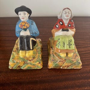 May include: A pair of vintage ceramic figurines depicting seated figures. One figure wears a blue top, black hat, and holds a bouquet. The other figure wears a red shawl and a white bonnet. Both figures are seated on a textured base with yellow, green, and brown tones.