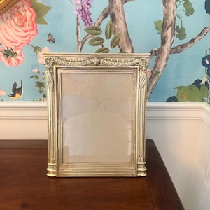 May include: A rectangular gold-toned picture frame featuring ornate neoclassical detailing, including a decorative frieze with carved swags and garlands along the top edge, vertical fluted columns on the sides, and a plain inner border.