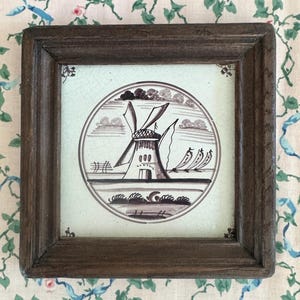 May include: A framed ceramic tile featuring a windmill design in shades of brown and white. The windmill is set within a circular frame against a pale green background. The tile is encased in a dark brown wooden frame, showcasing a rustic aesthetic.