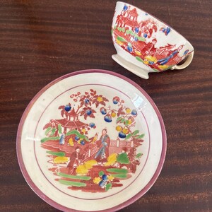 May include: A vintage teacup and saucer set with a detailed Asian-inspired design. The cup and saucer feature a red, white, and blue color scheme with illustrations of pagodas, figures, and foliage. The saucer has a pink rim.