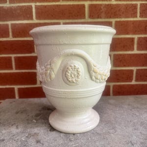 Puede incluir: Jarrón de cerámica blanquecina con borde ancho y base de pedestal. El jarrón presenta detalles decorativos en relieve, incluyendo un emblema central y diseños florales o de hojas. La superficie texturizada añade interés visual.