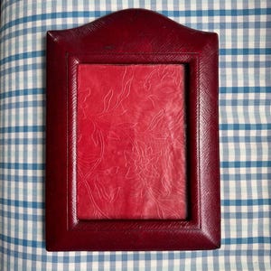 May include: A rectangular, burgundy-colored picture frame with a decorative top. The frame has a textured surface and surrounds a red background with a floral pattern. The frame is displayed on a blue and white checkered surface.