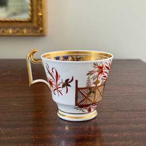 May include: A white teacup with gold trim and a gold handle. The cup features red and brown floral designs and a geometric pattern. The rim of the cup has a gold band. The teacup is on a wooden surface.
