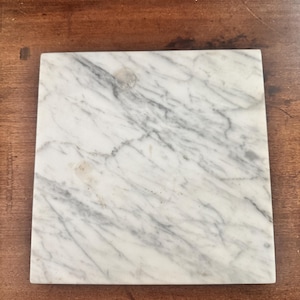 May include: Square marble coaster with grey veining on a white background. The coaster is made of natural stone and has a polished finish. The coaster is approximately 10 cm by 10 cm.