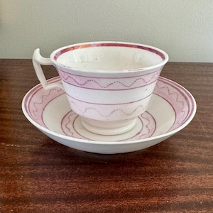 May include: A white teacup and saucer set with a pink and white design. The teacup has a handle and a pink rim. The saucer has a matching pink and white pattern. The set is placed on a dark wooden surface.