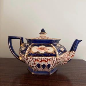 Könnte beinhalten: Eine blaue und weiße Teekanne mit goldenen Akzenten und einem floralen Design. Die Teekanne hat eine achteckige Form und einen Deckel mit einem Knopf.