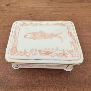 Puede incluir: Una caja rectangular de cerámica blanca con una ilustración de un pez en la tapa. La caja tiene un borde dorado y está decorada con ilustraciones de temática marina. La caja está soportada por cuatro pequeños pies.