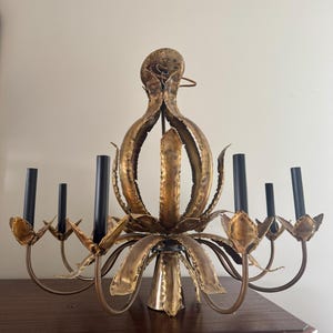 May include: A vintage gold-toned chandelier with a floral design. The chandelier has six black candle holders and a central light fixture. The fixture is made of metal and has a distressed finish, adding to its antique charm.