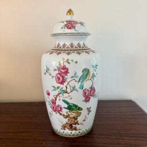May include: A white ceramic ginger jar with a lid, featuring a floral and bird design. The jar is adorned with pink flowers, green and blue birds, and brown branches. The lid is topped with a gold finial.