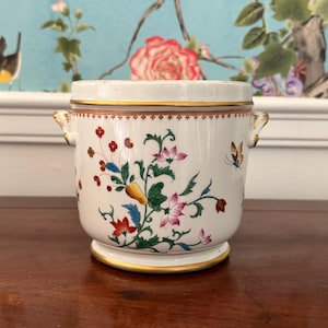 Puede incluir: Un recipiente de cerámica blanco con tapa, adornado con coloridos diseños florales y de mariposas. El recipiente tiene ribetes dorados y pequeñas asas. El fondo presenta un papel pintado floral.