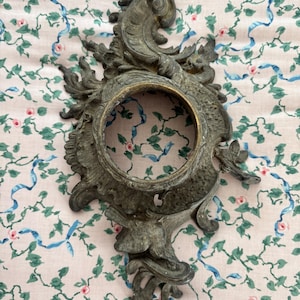 Op de afbeelding: Een sierlijke wandhouder of klokframe van donker metaal met ingewikkelde krulwerk- en acanthusbladmotieven. Het object heeft een centrale ronde opening met een gladde, messingkleurige binnenrand en een getextureerde, verouderde bronzen afwerking.