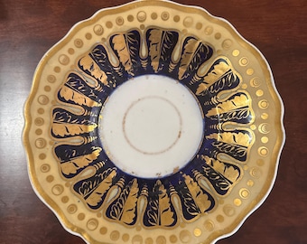 Puede incluir: Un platillo de porcelana vintage con borde festoneado. El platillo está decorado con un patrón floral azul y dorado.