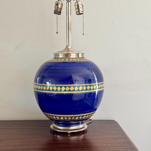 Op de afbeelding: Blauwe keramische tafellamp met een bolvormige voet en zilverkleurige metalen accenten. De voet heeft een band van gele en groene ruitpatronen. De lamp heeft twee fittingen en een zilverkleurige steel.