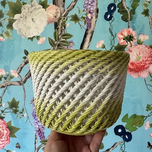 Puede incluir: Una maceta de cerámica con un diseño de cesta tejida y un degradado de color que va del verde lima en los bordes al blanco en el centro. La estructura consiste en hebras de cerámica diagonales que forman un patrón de celosía abierto.
