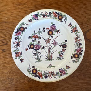 May include: A white ceramic plate with a floral design. The plate features a central arrangement of flowers in shades of pink, orange, and purple, surrounded by a border of similar floral patterns. The plate is set against a wooden surface.