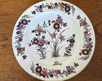 Op de afbeelding: Een witte keramische plaat met een bloemenontwerp. De plaat heeft een centrale opstelling van bloemen in tinten roze, oranje en paars, omgeven door een rand van vergelijkbare bloemmotieven. De plaat staat op een houten oppervlak.